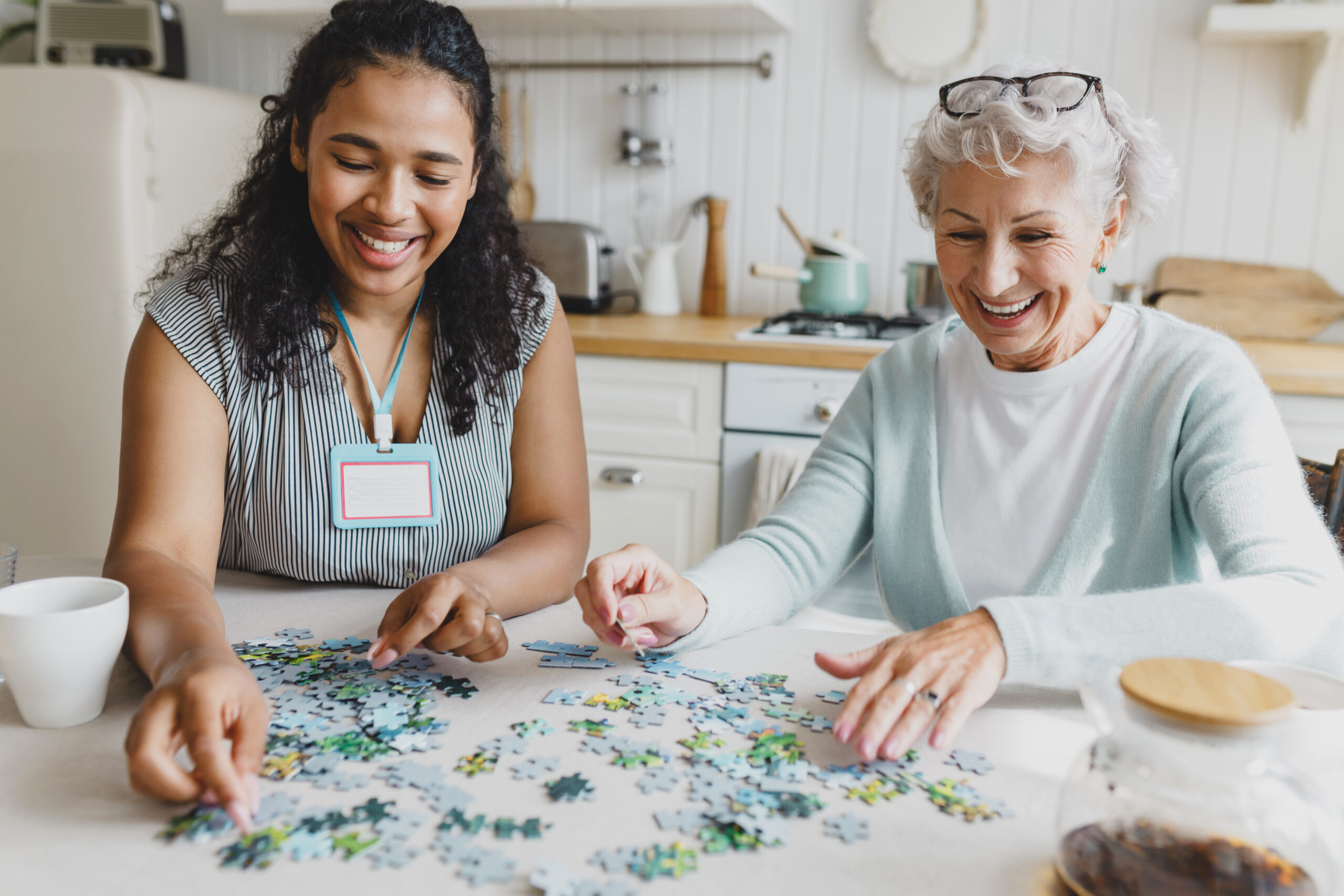 Portrait of african american female social worker volunteer playing puzzle game with cheerful senior lady, having fun, laughing searching for right piece sitting together at kitchen table Female caregiver playing puzzle game with cheerful senior lady at a kitchen table